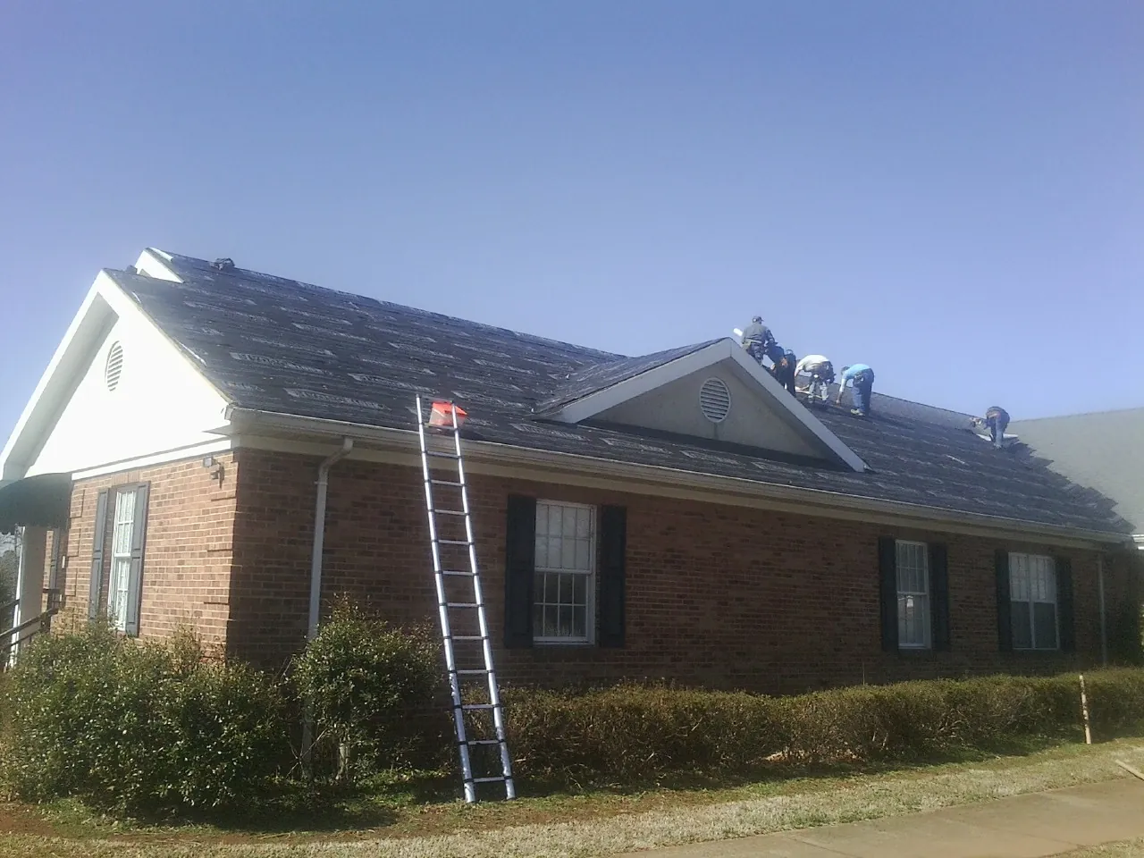 Crew tearing shingles off a brick ranch roof with a ladder set — Emergency Roof Repair work by our crew in Cardington