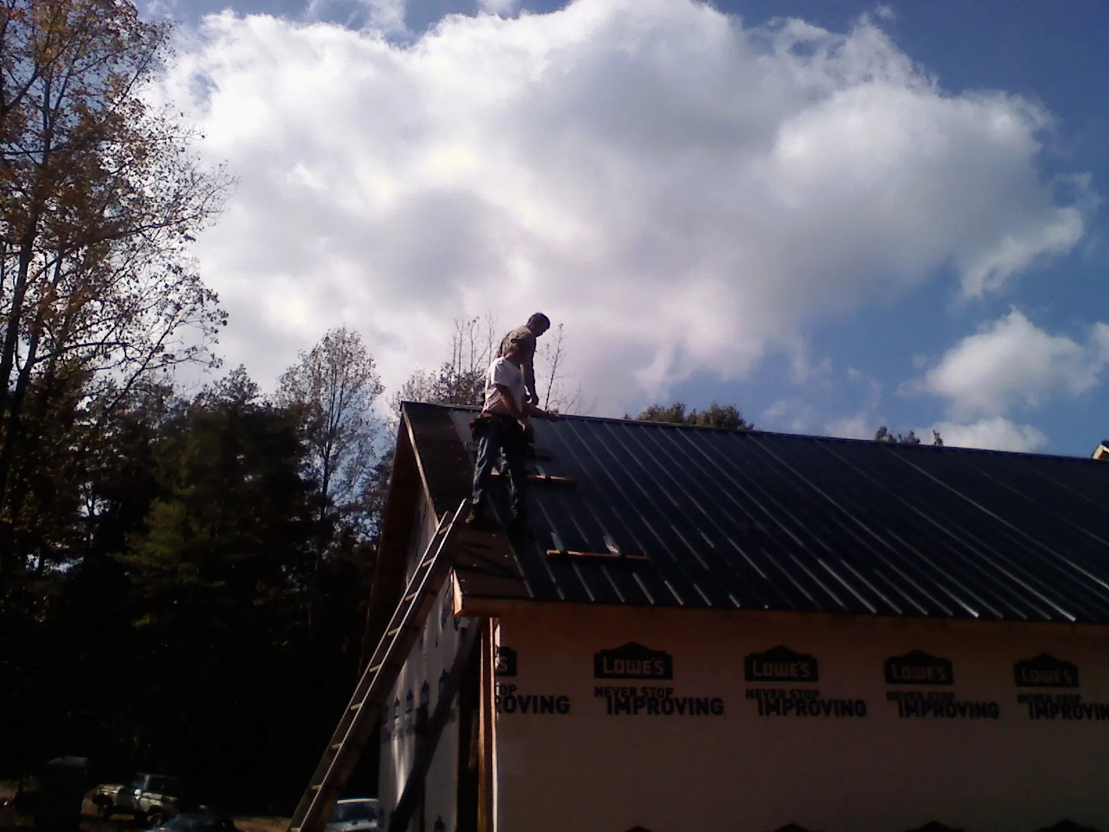 Roofer fastening dark metal panels high against a cloudy sky, from a Roof Inspection job in Cardington